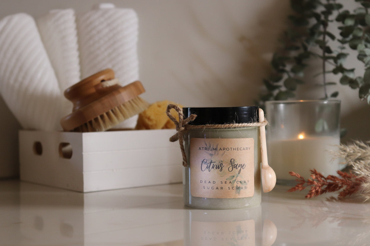 A jar of Sugar Scrub- Citrus Sage with a label and a small wooden spoon, placed on a counter next to a candle and a bath brush.