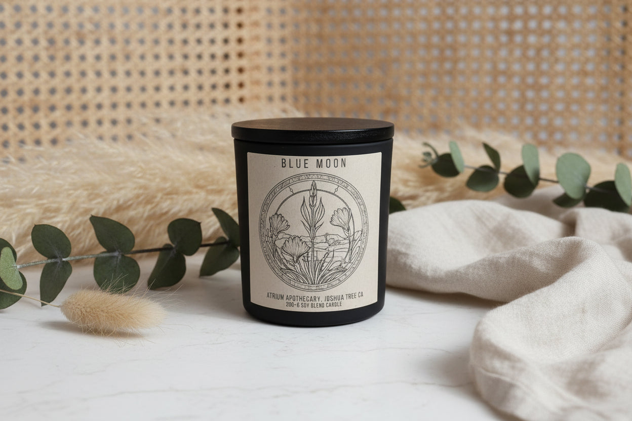 A black candle jar with a label that reads 'Blue Moon Atrium Apothecary, Joshua Tree CA', featuring an illustration of plants and flowers.
