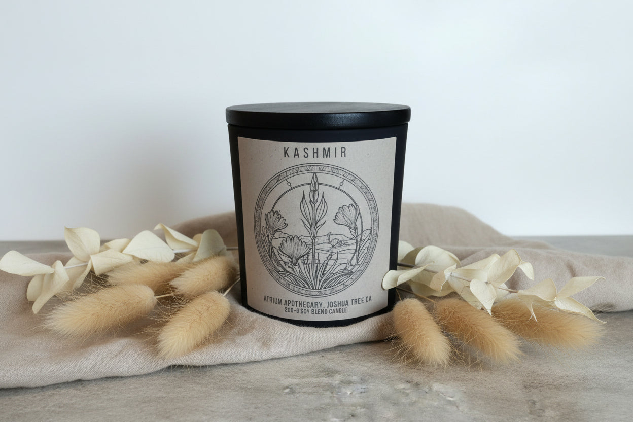 A matte black jar candle with a wooden lid, labeled 'KASHMIR' on the front. The label also features a drawn circular design with evergreen plants and a floral motif.