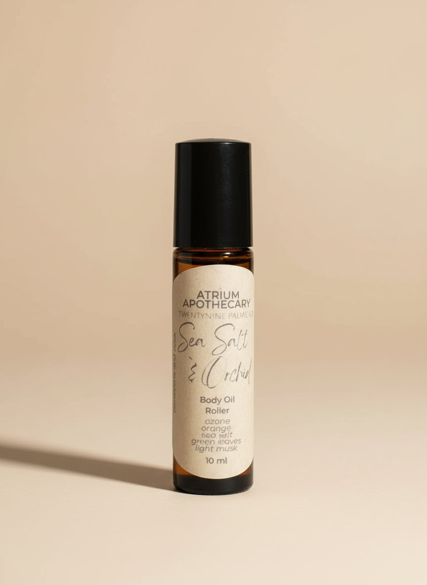 Body oil roller bottle labeled 'Atrium Apothecary' on a surface with a plant in the background